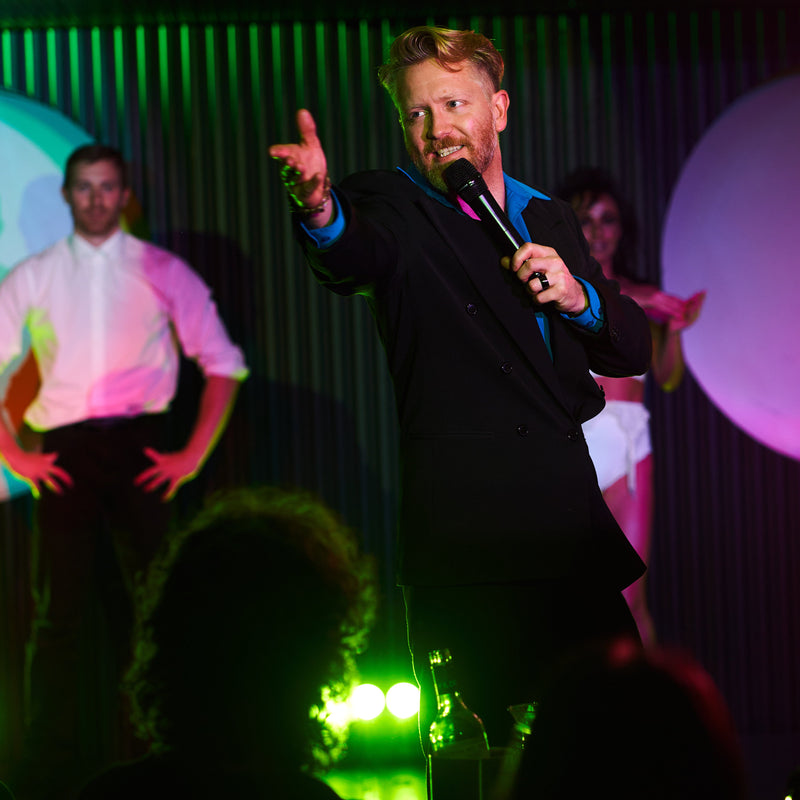 Comedian MC with a microphone does a dramatic gesture on stage while other cabaret performers stand behind him in vaudeville dinner show in Potts Point